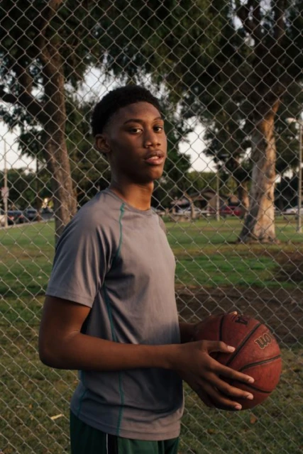 Young man in athletic wear holding a basketball outdoors in park setting, portrait shot against chain link fence, looking toward the camera.