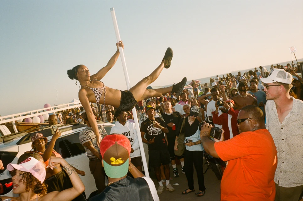 Woman in bikini performs on a pole at an outdoor party on a sunny day, with a large crowd of people gathered around to watch.