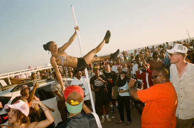 Woman in bikini performs on a pole at an outdoor party on a sunny day, with a large crowd of people gathered around to watch.