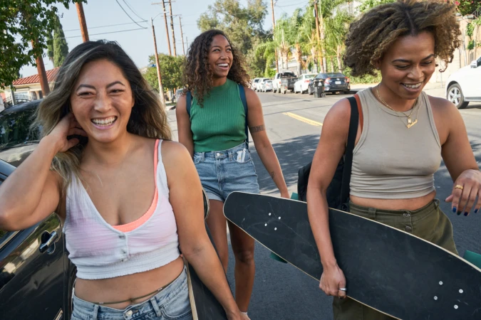 Three young women walk on a sunny street with skateboards, smiling and laughing, with houses and cars in the background.