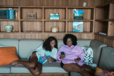 Two Black women relaxing together on a light grey sectional sofa in a wood-paneled room while looking at a smartphone.