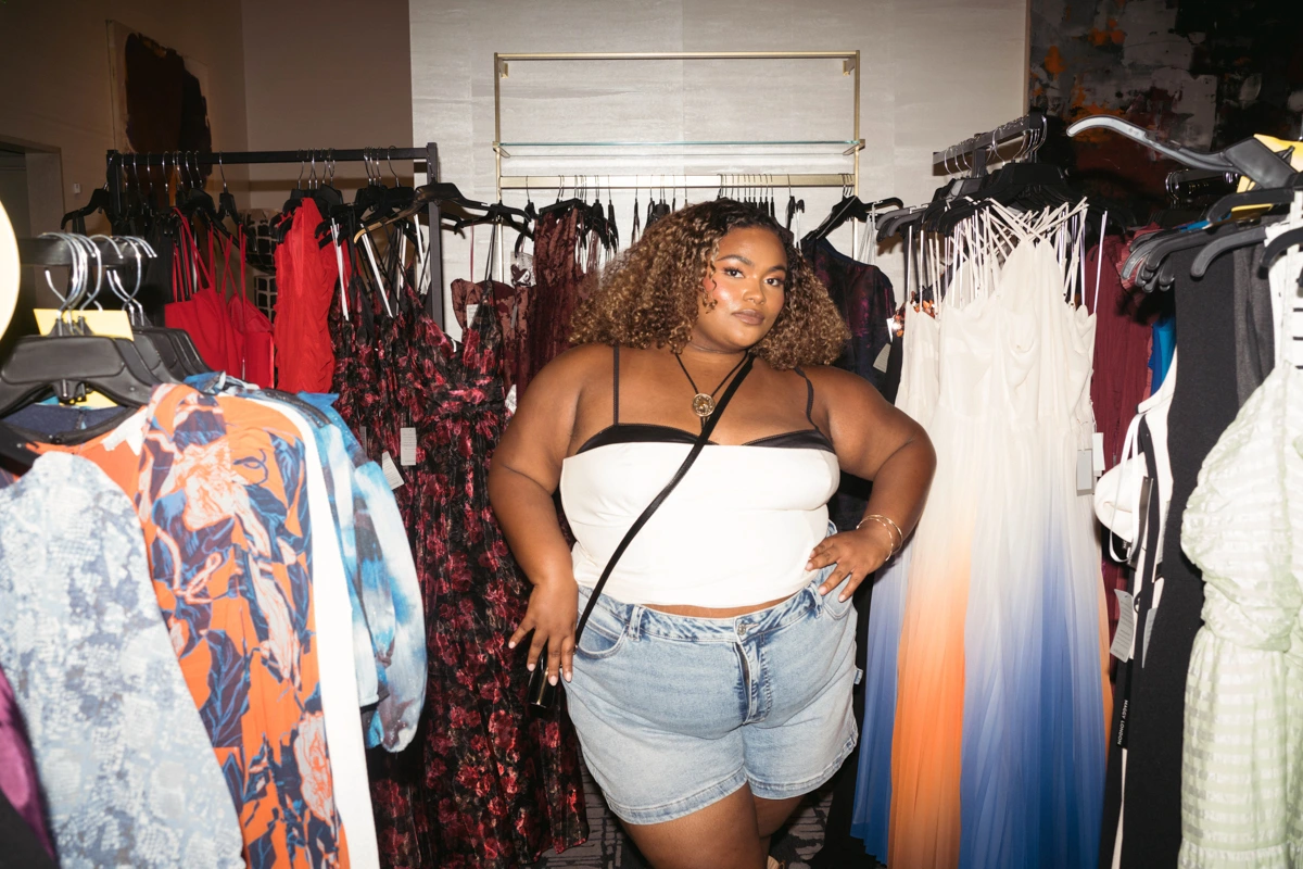 Plus size woman in clothing store wearing white top and denim shorts surrounded by racks of dresses in Los Angeles.
