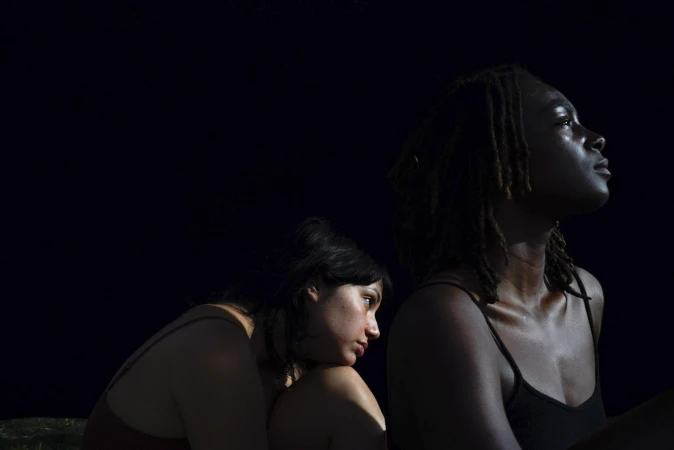 Two women posing against a dark background in low-key lighting by photographer Gabriella Angotti-Jones.