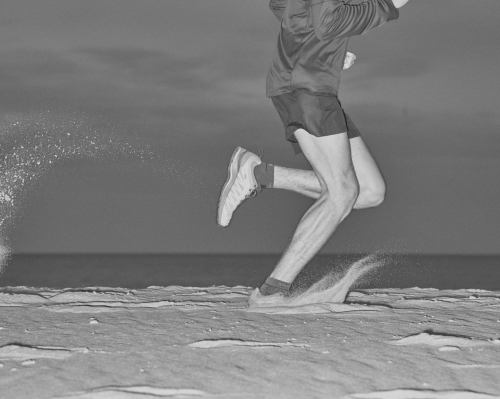 Black and white image of a man running on a sandy surface with the ocean in the background, captured in a medium shot.