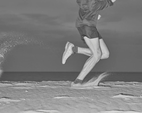 Black and white image of a man running on a sandy surface with the ocean in the background, captured in a medium shot.