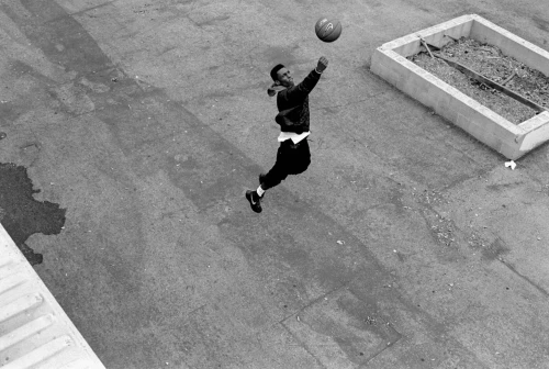 High-angle shot of a man in mid-air, reaching for a basketball on a paved surface with a concrete planter in the background, captured in black and white.