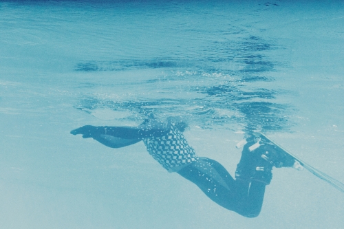 Underwater view of a person water skiing with a checkered swimsuit in clear, blue water with sunlight reflecting on the surface.