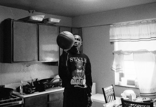A young adult male spins a basketball on his finger in a kitchen, wearing a graphic sweatshirt with a window in the background.