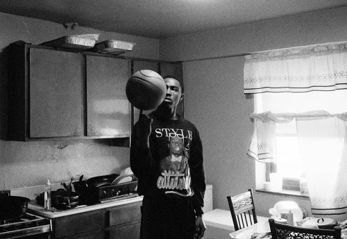 A young adult male spins a basketball on his finger in a kitchen, wearing a graphic sweatshirt with a window in the background.