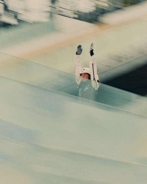 A person in a white jacket and helmet rides down an icy track with their arms up, suggesting speed and motion in a winter sport.