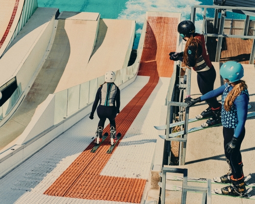 Three women in sports gear at the top of an indoor ski slope, ready to ski. One skier heads down slope. Bright, artificial light.