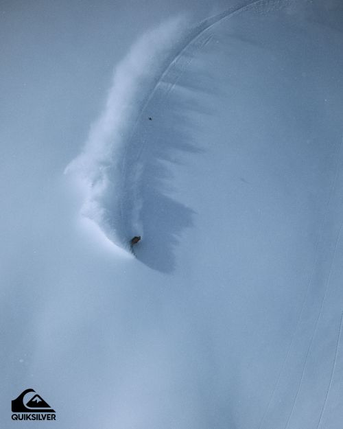An aerial shot captures a snowboarder kicking up powder while carving through a vast, snow-covered landscape under soft lighting.