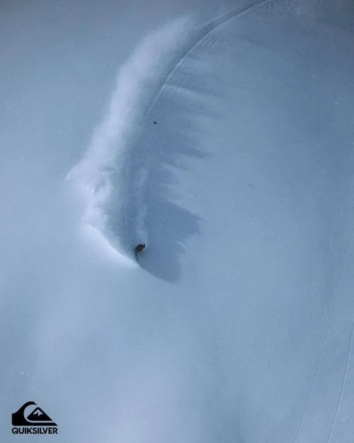 An aerial shot captures a snowboarder kicking up powder while carving through a vast, snow-covered landscape under soft lighting.