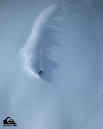 An aerial shot captures a snowboarder kicking up powder while carving through a vast, snow-covered landscape under soft lighting.