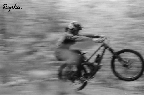 A person rides a mountain bike on a dirt trail surrounded by trees in a black-and-white action shot with motion blur.
