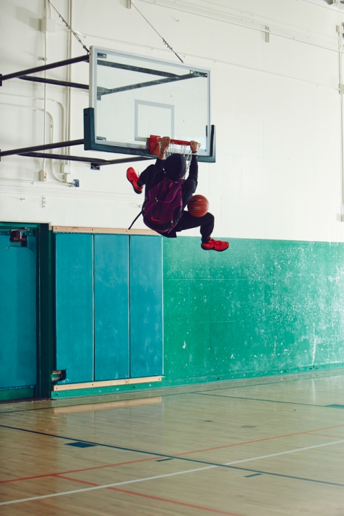 An athlete hangs from a basketball hoop with a ball, wearing red sneakers and backpack indoors, spotlighting sport and physical activity.