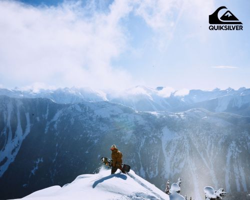 Snowboarder stands on a snowy mountaintop with a board, overlooking a vast mountain range under a bright, cloudy sky.