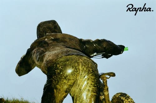 Man and mountain bike covered in mud, shot from behind against a light blue sky, with the Rapha logo visible in the top right corner.