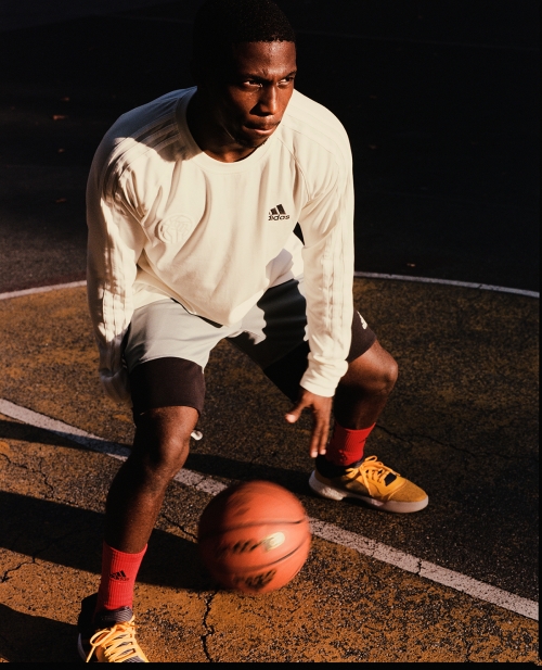 Young Black man dribbling a basketball on an outdoor court, wearing a white long-sleeve shirt, grey shorts, and orange socks and sneakers.