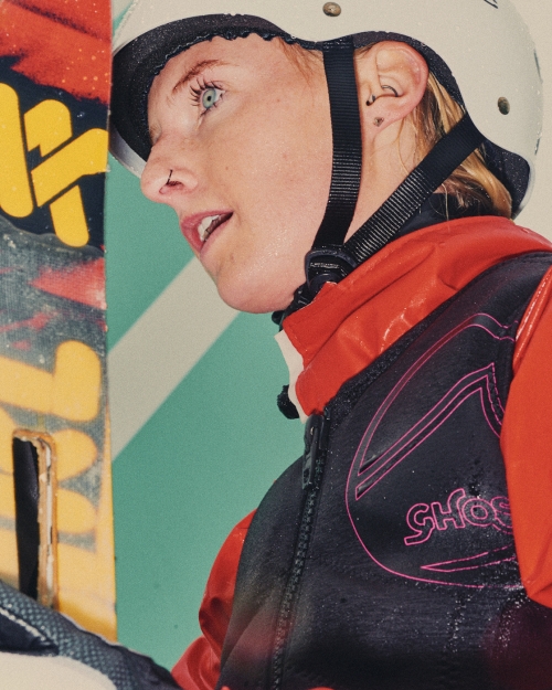 Close-up of a blonde-haired, blue-eyed young adult in a white helmet and black and red vest, looking to the left in a bright indoor setting.