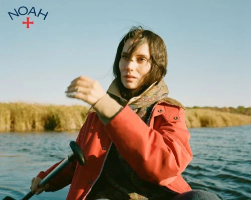 Woman in red coat paddles on a river near grassy shore, wearing a patterned scarf under a blue sky, logo of Noah upper left.