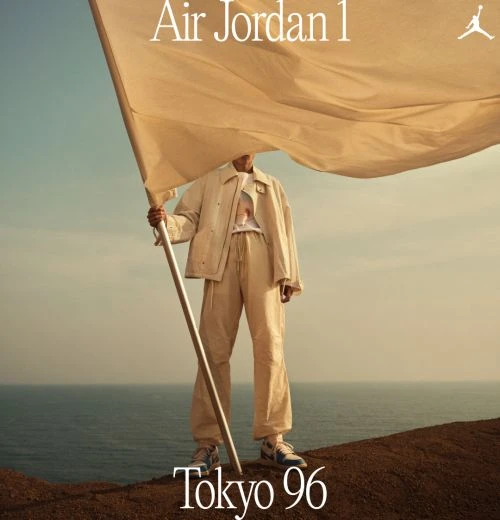 An adult wearing beige outfit holds a flag on a hill near the ocean. 'Air Jordan 1' and 'Tokyo 96' text visible in the shot.
