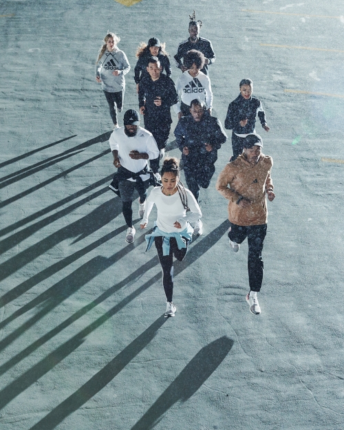 Group of diverse runners in athletic wear sprinting outdoors on gray pavement in overcast conditions, creating long shadows.