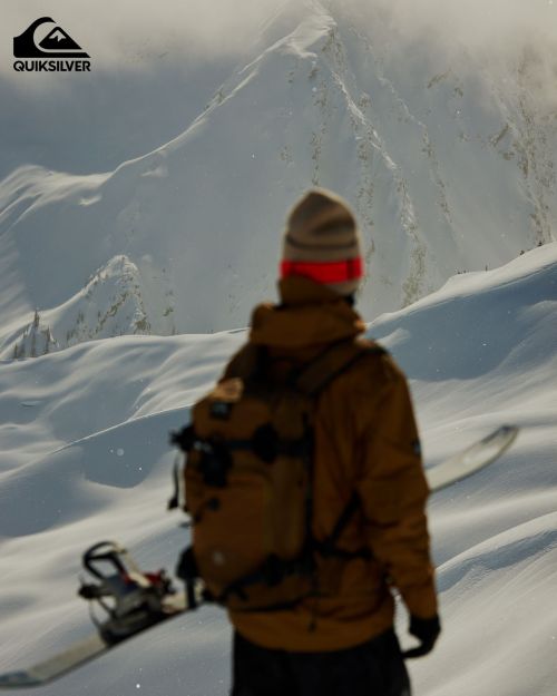 Person with snowboard and backpack in snow covered mountain landscape wearing winter hat and tan jacket under overcast lighting.