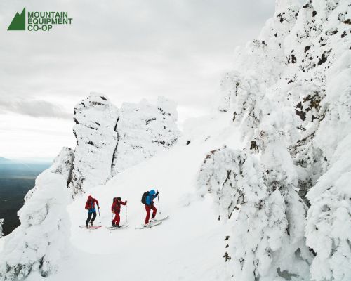 Three people are cross-country skiing up a snow-covered mountain with frosted trees and rock formations on a cloudy day in winter.