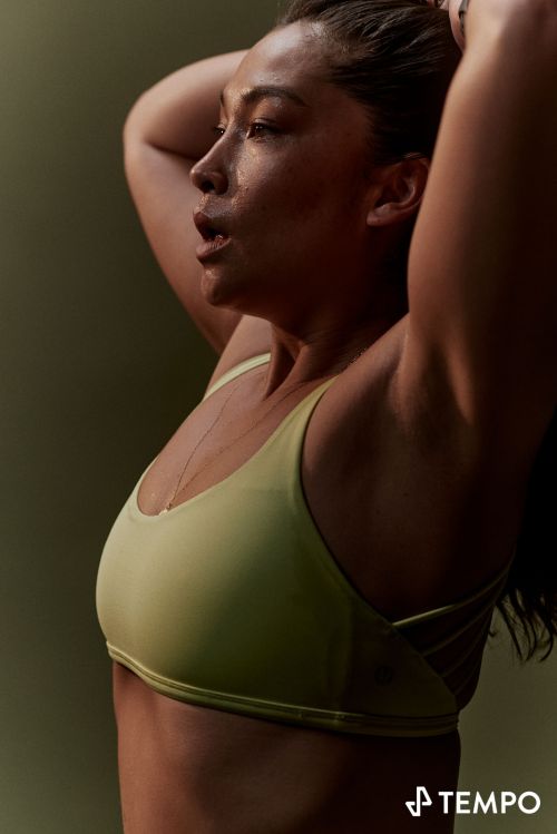 Side-view of an adult female with dark hair and olive skin, wearing a sports bra and holding her hair up.