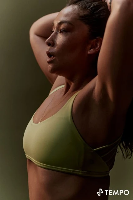 Side-view of an adult female with dark hair and olive skin, wearing a sports bra and holding her hair up.