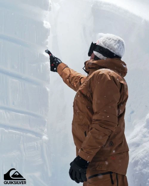 Man in a brown jacket and snow goggles is outside near a wall of snow. He's wearing a knit cap and gloves, inspecting the snow.