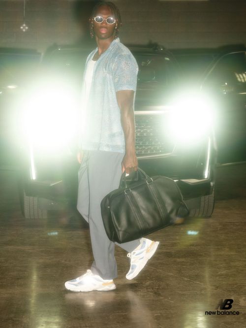 A male model poses in front of a car in a dim parking garage, wearing sunglasses, a blue shirt, grey pants, New Balance sneakers, and holding a black bag.