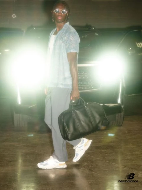 A male model poses in front of a car in a dim parking garage, wearing sunglasses, a blue shirt, grey pants, New Balance sneakers, and holding a black bag.