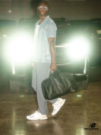 A male model poses in front of a car in a dim parking garage, wearing sunglasses, a blue shirt, grey pants, New Balance sneakers, and holding a black bag.
