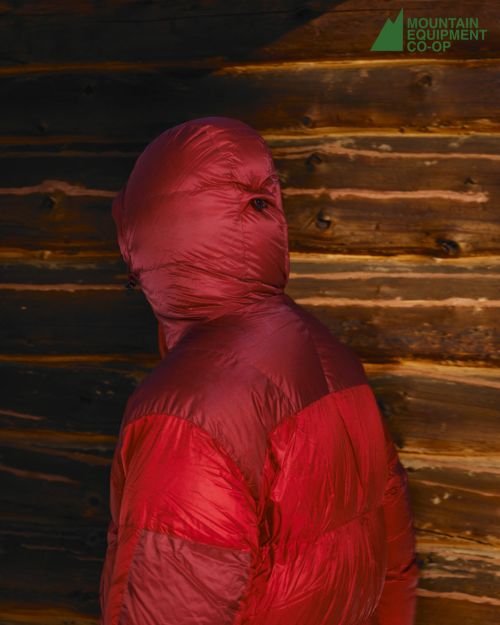 Person in a red puffer jacket with hood pulled over head standing in front of a textured wooden wall. Mountain Equipment Co-op logo.