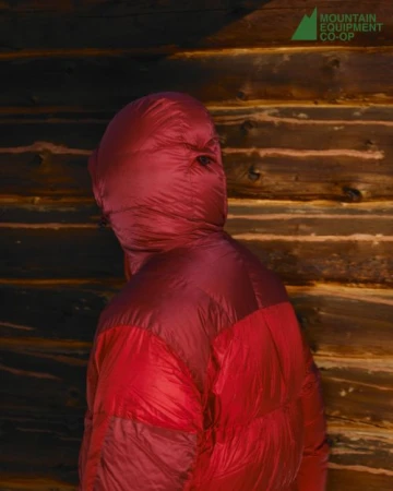 Person in a red puffer jacket with hood pulled over head standing in front of a textured wooden wall. Mountain Equipment Co-op logo.