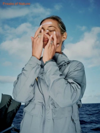 Woman wearing a grey windbreaker applying sunscreen to her face outdoors with ocean and sky in background