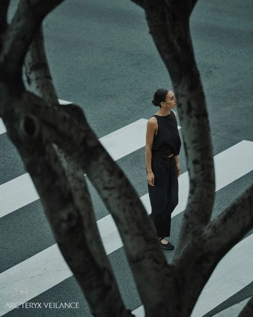 A woman in black apparel crossing an urban crosswalk is partially framed by tree branches, shot in a high-angle composition, cool lighting.