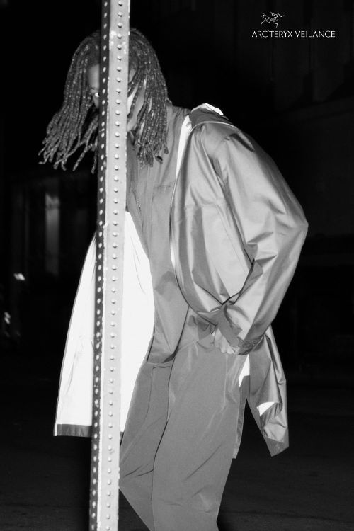 Black and white portrait of a Black adult male with dreadlocks standing next to a striped pole wearing a jacket and pants outdoors.