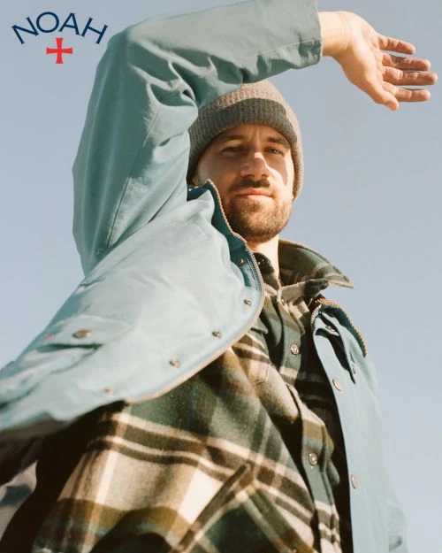Adult male shields eyes from sun in medium shot, wearing a beanie, plaid shirt, and light blue jacket against a light blue sky.