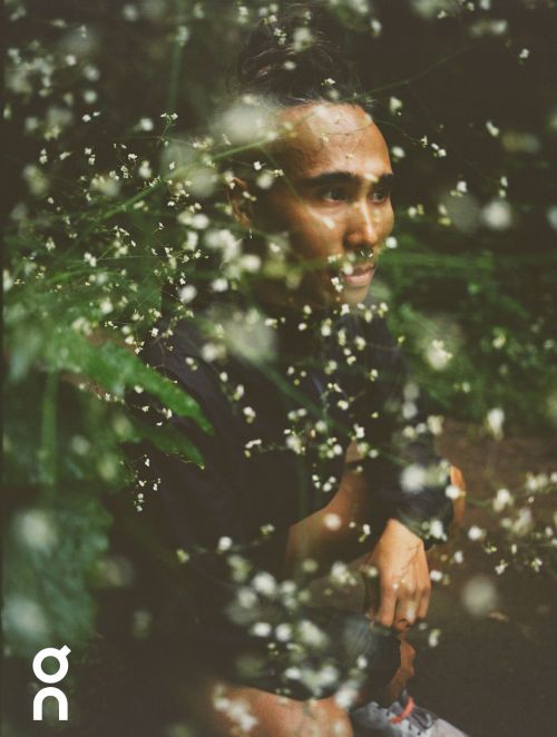 Close-up portrait of a man with a top knot, looking to the right, surrounded by white flowers and green foliage in an outdoor setting.