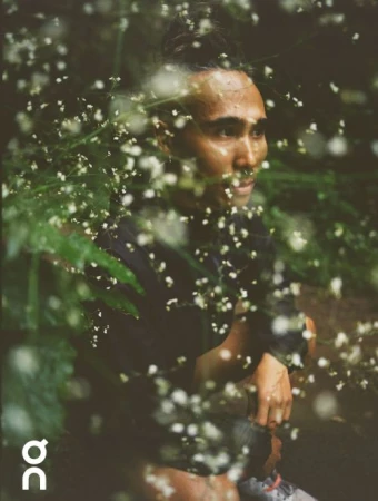 Close-up portrait of a man with a top knot, looking to the right, surrounded by white flowers and green foliage in an outdoor setting.