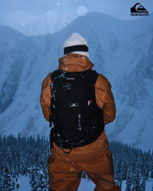 Man in a snowsuit and backpack stands looking out at snow-covered mountain landscape, winter forest visible below, Quiksilver logo in corner.