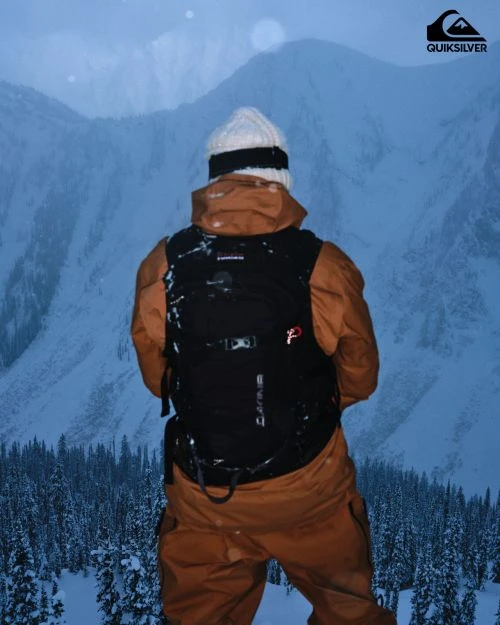 Man in a snowsuit and backpack stands looking out at snow-covered mountain landscape, winter forest visible below, Quiksilver logo in corner.