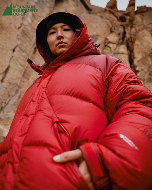 Eye-level shot of an Asian woman in a red hooded puffer jacket against a rock formation backdrop, captured with a shallow depth of field.