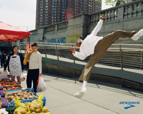 Adult male in mid-air, doing a split jump on an urban sidewalk with a street market, high-rise buildings, and the Brooks logo at the bottom.