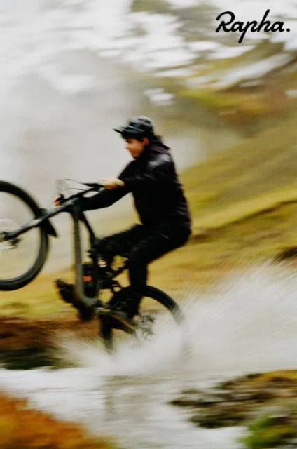 An adult man in black sportswear rides a mountain bike through shallow water with a blurred background of grass and cloudy sky.
