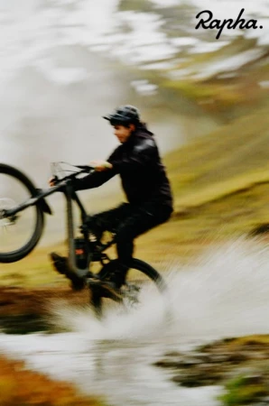An adult man in black sportswear rides a mountain bike through shallow water with a blurred background of grass and cloudy sky.