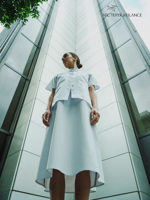 Woman in white architectural clothing posing in front of a modern high-rise building with glass and metal facade, shot from a low angle.